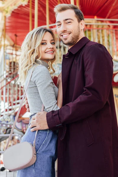 Sourire couple élégant câlin près du carrousel dans le parc d'attractions et en regardant la caméra — Photo de stock