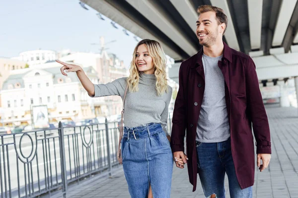 Fidanzata sorridente in abito autunnale che punta su qualcosa al fidanzato mentre cammina sotto il ponte in città — Foto stock