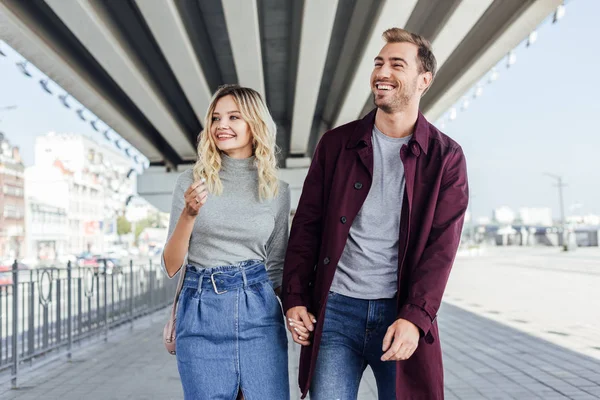 Romantic smiling couple in autumn outfit holding hands and walking under bridge in city — Stock Photo
