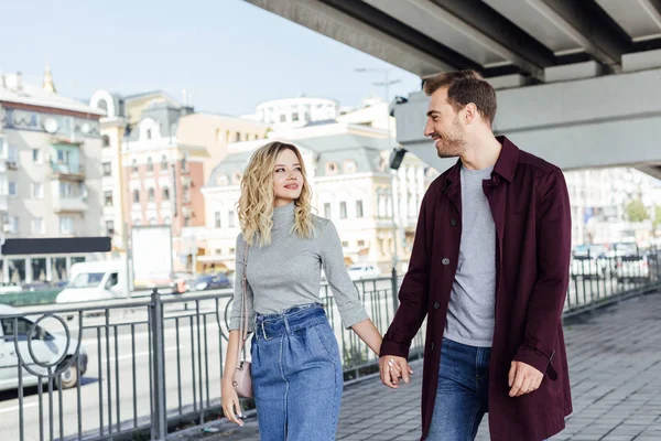 Pareja romántica en traje de otoño tomados de la mano y mirándose unos a otros bajo el puente en la ciudad - foto de stock