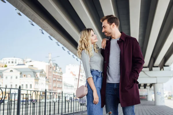 Tender couple in autumn outfit looking at each other under bridge in city — Stock Photo