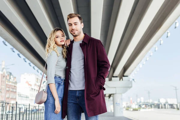 Stilvolles Paar im Herbst-Outfit umarmt sich unter Brücke in der Stadt und blickt in die Kamera — Stockfoto