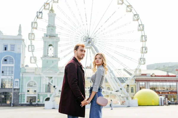 Vista lateral do casal em roupa de outono de mãos dadas com roda gigante no fundo — Fotografia de Stock