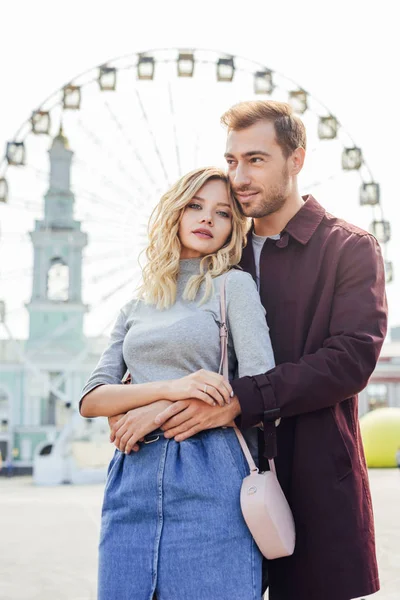 Pareja joven en traje de otoño abrazándose con rueda de observación en el fondo - foto de stock