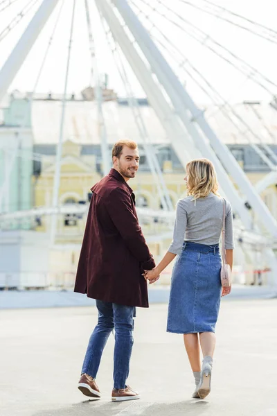 Rückansicht eines Paares im Herbst-Outfit, das Händchen hält und zum Beobachtungsrad läuft — Stockfoto