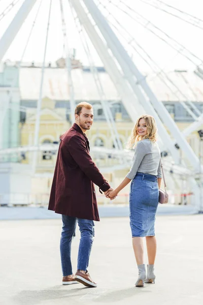 Vista trasera de la pareja sonriente en traje de otoño tomados de la mano y caminando a la rueda de los hurones - foto de stock