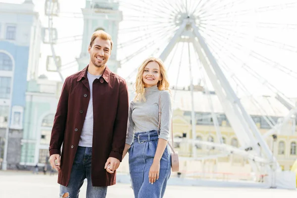 Happy couple in autumn outfit holding hands and standing near observation wheel — Stock Photo