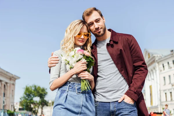 Vista de ángulo bajo de la pareja en traje de otoño abrazándose en la calle en la ciudad, novia sosteniendo ramo de rosas — Stock Photo