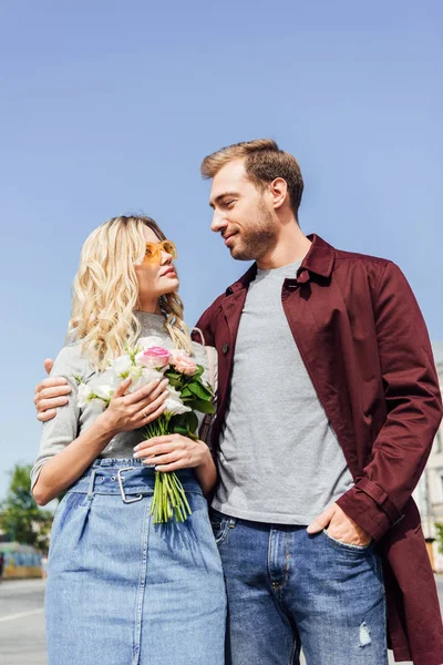 Vista de ángulo bajo de pareja en traje de otoño mirándose en la calle, novia sosteniendo ramo de rosas - foto de stock