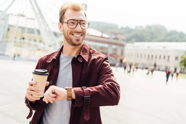 Bell'uomo sorridente in abito autunnale che tiene la tazza di caffè usa e getta e controlla il tempo in città — Foto stock