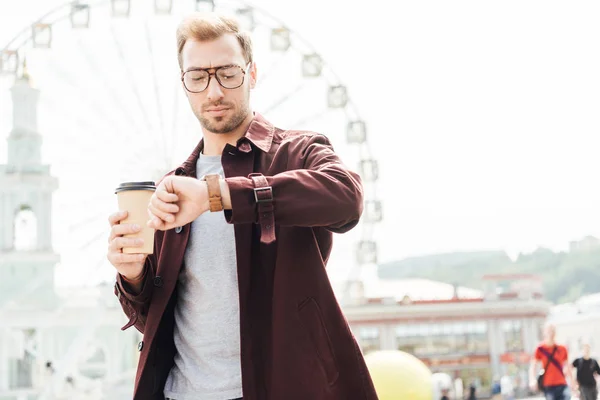 Vue à angle bas du bel homme en tenue d'automne tenant le café dans une tasse en papier et vérifiant l'heure près de la roue d'observation — Photo de stock