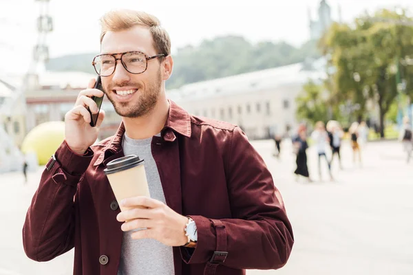 Улыбающийся мужчина в осеннем наряде держит одноразовую чашку кофе и разговаривает по смартфону в городе — стоковое фото