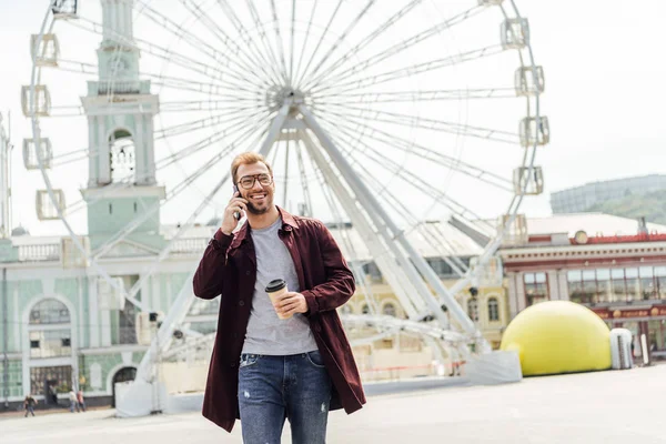 Uomo sorridente in abito autunnale in possesso di caffè per andare e parlare con smartphone vicino ruota di osservazione — Foto stock