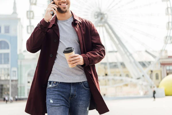 Cropped image of man in autumn outfit holding coffee to go and talking by smartphone near observation wheel — Stock Photo