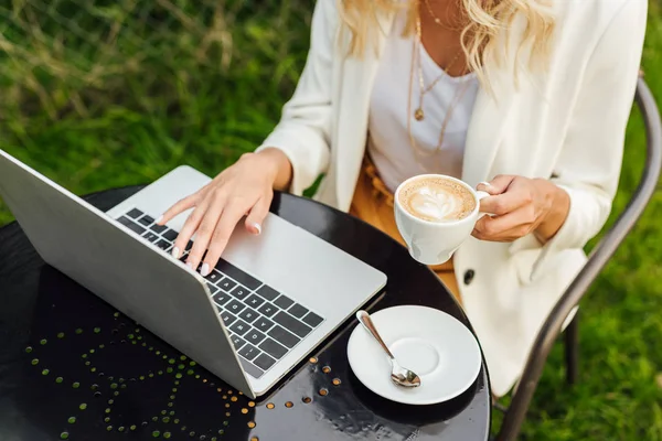 Обрізане зображення жінки за допомогою ноутбука і тримає чашку кави за столом в саду — стокове фото