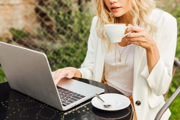 Image recadrée de la femme en utilisant un ordinateur portable et tenant une tasse de café à table dans le jardin — Photo de stock