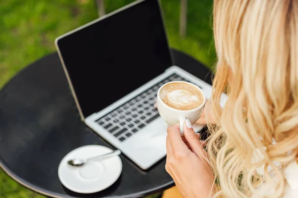 Обрезанное изображение женщины с чашкой кофе, ноутбук с пустым экраном на столе в саду — стоковое фото