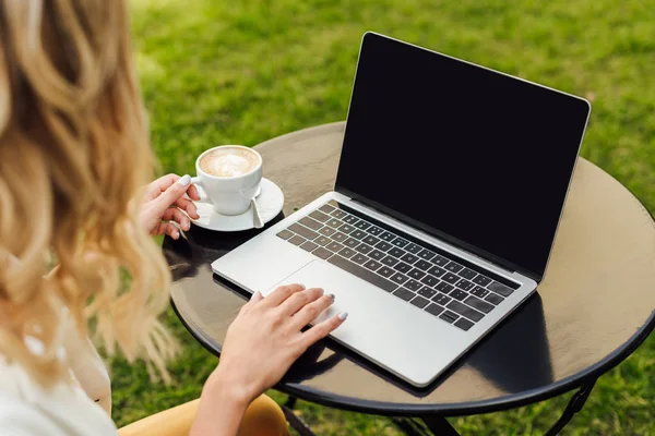 Image recadrée de la femme en utilisant un ordinateur portable avec écran vierge et tenant une tasse de café sur la table dans le jardin — Photo de stock