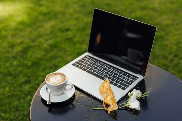 Laptop, cup of coffee, sunglasses and flowers on table in garden — Stock Photo