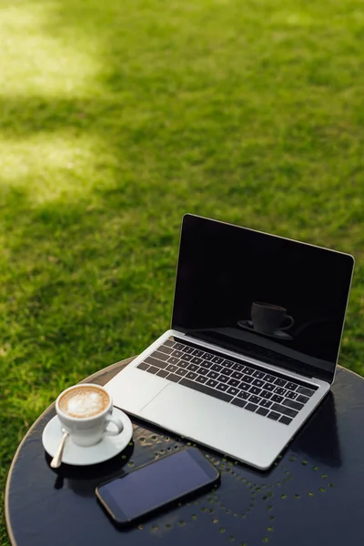 Portatile e smartphone con schermo bianco e tazza di cappuccino sul tavolo in giardino — Foto stock