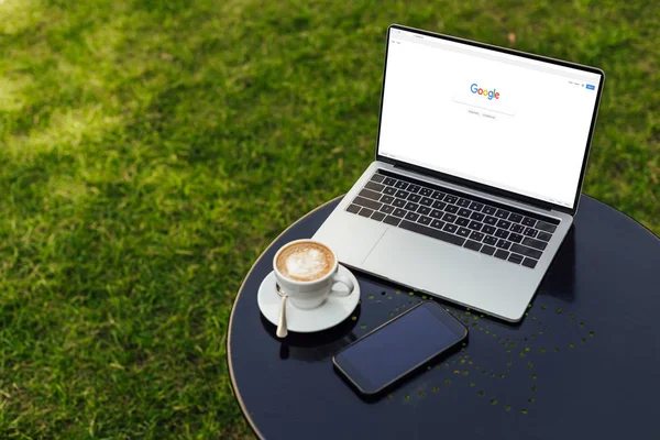 Ноутбук с загруженной страницей Google, чашка кофе и смартфон на столе в саду — стоковое фото