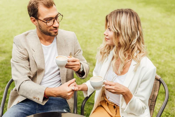 Smiling couple in autumn outfit sitting on chairs in cafe, holding hands and looking at each other — Stock Photo