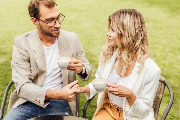 Pareja sonriente en traje de otoño sentado en sillas en la cafetería, tomados de la mano y mirándose el uno al otro - foto de stock