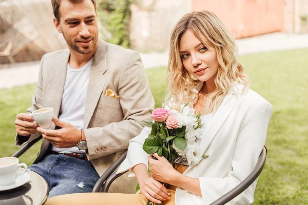 Attractive girlfriend holding bouquet of flowers in cafe, boyfriend holding cup of coffee — Stock Photo