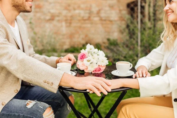 Imagen recortada de pareja en traje de otoño tomados de la mano en la mesa en la cafetería - foto de stock