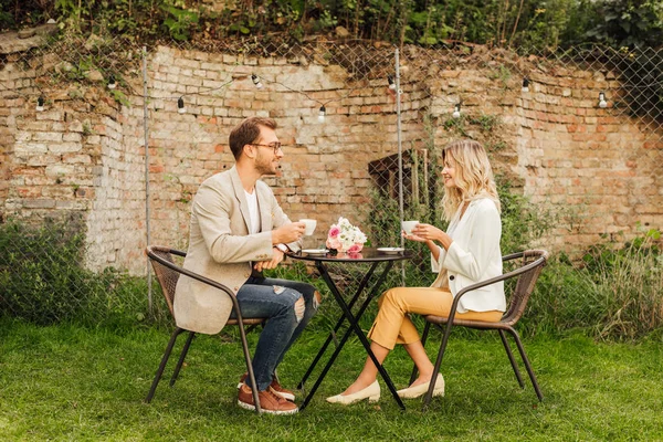 Боковой вид на пару, сидящую за столом в кафе с чашками кофе и букетом на столе — стоковое фото