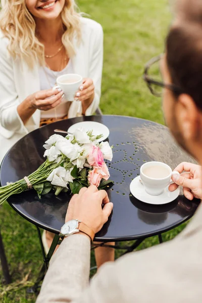 Immagine ritagliata di coppia in abito autunnale seduto a tavola in caffè con tazze di caffè e bouquet sul tavolo — Foto stock