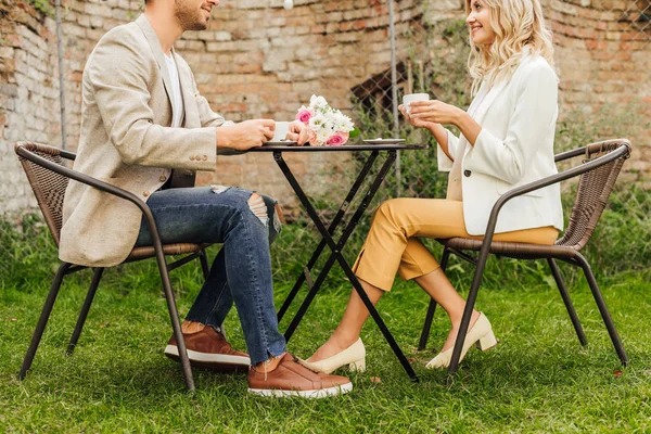 Vue latérale du couple en tenue d'automne assis à table dans un café avec des tasses de café et un bouquet sur la table — Photo de stock