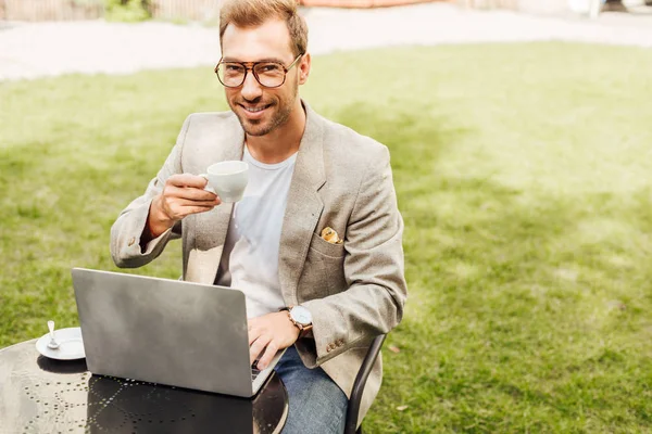 Smiling freelancer in autumn outfit working with laptop at table in city and holding cup of coffee — Stock Photo