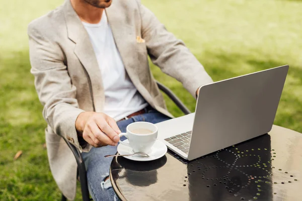 Imagen recortada de freelancer en traje de otoño sentado en la mesa con ordenador portátil y taza de café en el jardín - foto de stock
