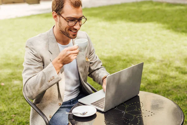 Bel freelance in abito autunno lavorando con il computer portatile a tavola in città e tenendo in mano una tazza di caffè — Foto stock