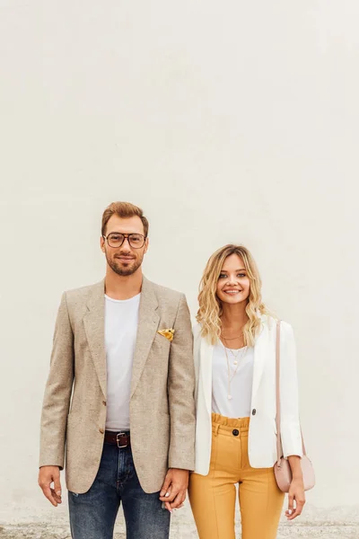 Smiling couple in autumn outfit standing near beige wall on street and looking at camera — Stock Photo