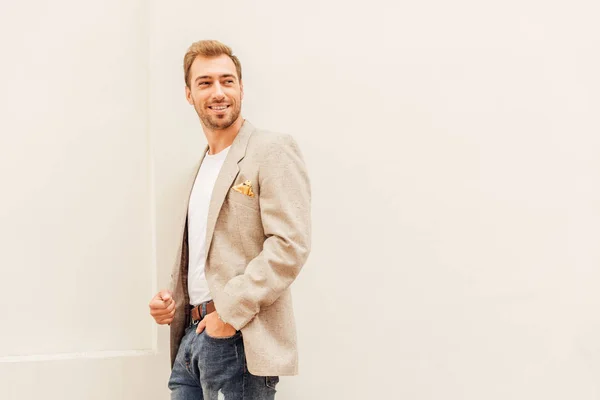 Smiling handsome man in jacket and jeans standing near beige wall in city — Stock Photo