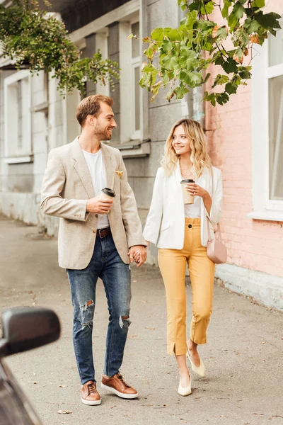 Couple gai en tenue d'automne tenant la main, se regardant et marchant dans la rue avec des tasses à café jetables — Photo de stock