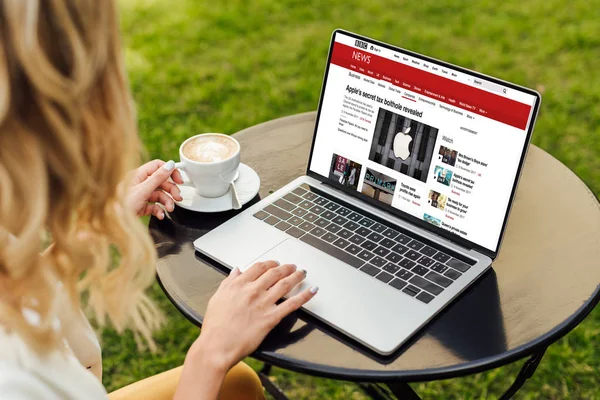 Imagen recortada de la mujer usando el ordenador portátil con la página de noticias de la penes cargados en la mesa en el jardín - foto de stock