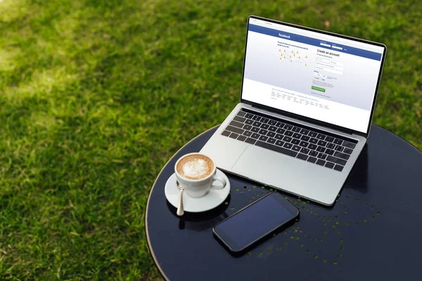 Laptop with loaded facebook page, cup of cappuccino and smartphone on table in garden — Stock Photo