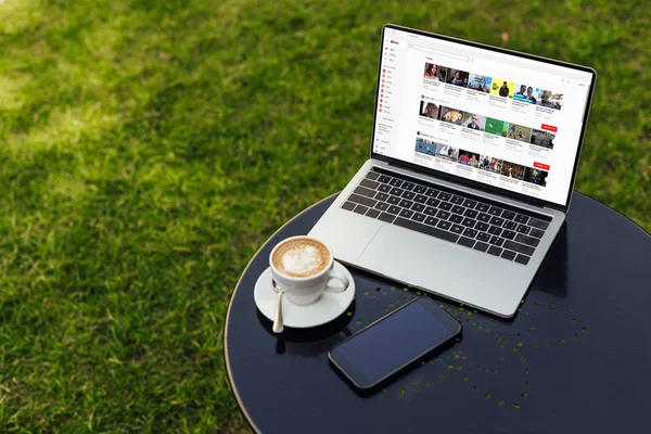 Laptop with loaded youtube page, cup of cappuccino and smartphone on table in garden — Stock Photo