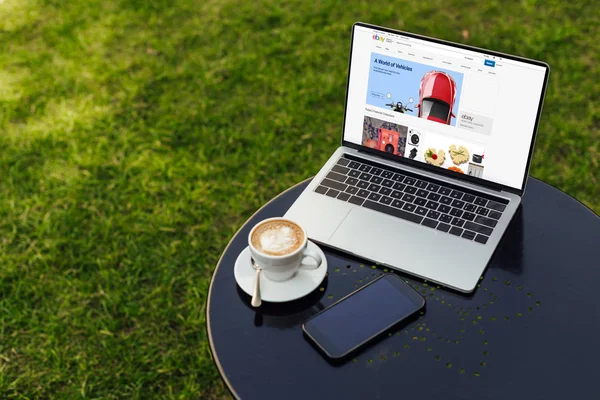 Portátil con página ebay cargado, taza de capuchino y teléfono inteligente en la mesa en el jardín - foto de stock