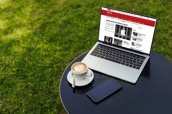 Laptop mit geladener bbc-Nachrichtenseite und Smartphone auf Tisch im Garten — Stockfoto