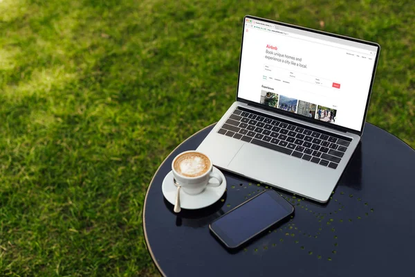 Laptop with loaded airbnb page, cup of coffee and smartphone with blank screen on table in garden — Stock Photo