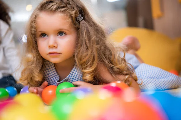 Adorável criança deitada no tapete com bolas coloridas no jardim de infância e olhando para longe — Fotografia de Stock