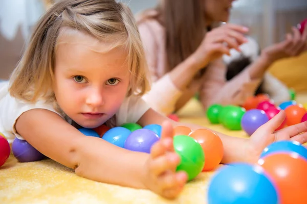 Criança adorável deitado no chão com brinquedos coloridos e olhando para a câmera no jardim de infância — Fotografia de Stock