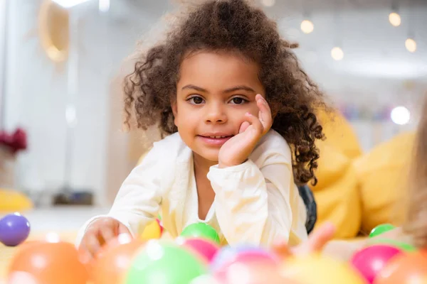 Sorridente bambino africano americano sdraiato su tappeto con giocattoli e guardando la fotocamera nella scuola materna — Foto stock