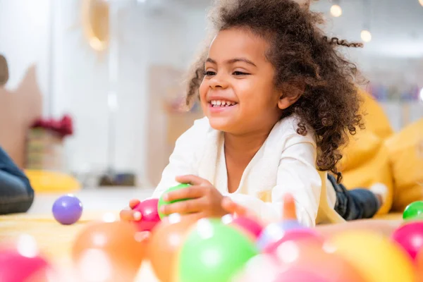 Fröhliches afrikanisch-amerikanisches Kind liegt mit Spielzeug auf Teppich und schaut im Kindergarten weg — Stockfoto