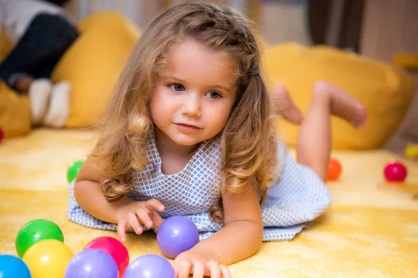 Adorable niño caucásico acostado en la alfombra con bolas de colores y mirando hacia otro lado en el jardín de infantes - foto de stock