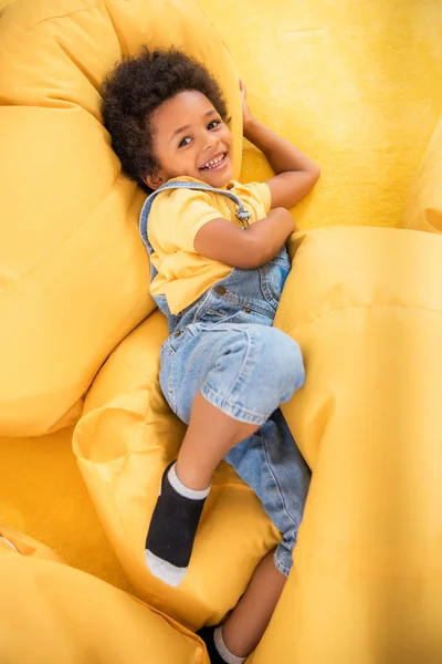 Visão de alto ângulo do menino afro-americano sorridente deitado em cadeiras de saco de feijão e olhando para a câmera no jardim de infância — Fotografia de Stock